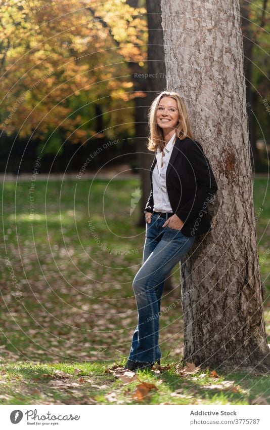 Smiling woman near tree in park stroll relax enjoy season sunlight garden carefree female autumn fall style recreation city casual rest trendy stand lady lean