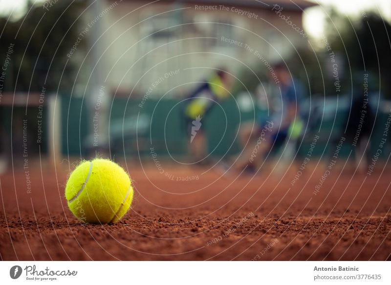 Closeup of a bright yellow tennis ball standing in a clay ground playing field. Outlines of two people in the background, tennis players taking a break during a championship