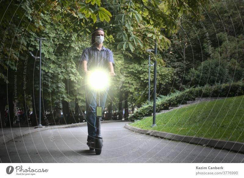 Masked young man riding eco friendly electric scooter with headlights on at park ride transportation technology electronic wheels feet people city green road