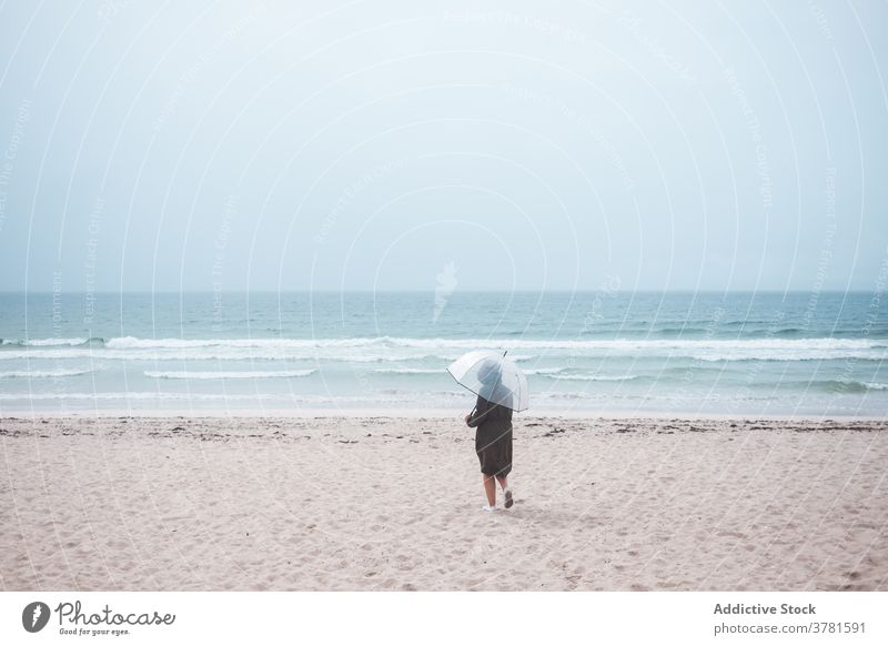 Anonymous person with umbrella walking on sandy beach sea alone travel ocean lonely stormy overcast traveler shore coast nature wave weather tourism seaside