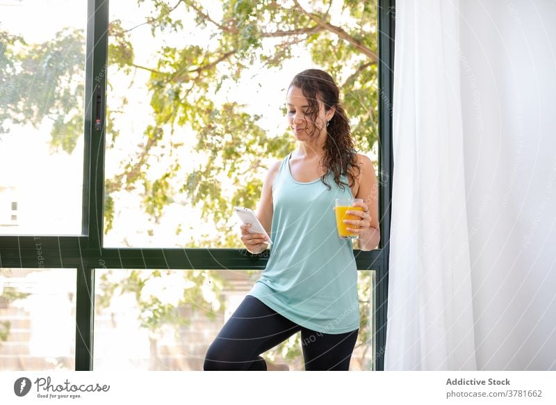 Calm woman with juice using smartphone at home morning orange enjoy chat social media weekend female fresh glass window domestic drink beverage device gadget