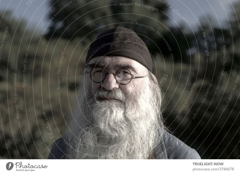 Klaus Hugler, deacon and religious educator, following in the footsteps of Lev Nikolaevich Count Tolstoy, here with a dark cap, small glasses and a powerful, glowing beard when looking into the camera in front of the bushes and trees of Babelsberg Park