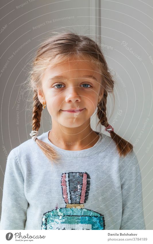 Close up shot of beautiful сaucasian little girl with pigtails wearing cozy sweater, looking at camera with charming smile, posing against light space of concrete wall. Concept of happy childhood