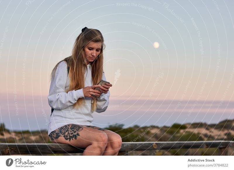 Peaceful woman browsing smartphone on promenade at sunset travel using twilight relax embankment female fence wooden surfing traveler calm internet vacation