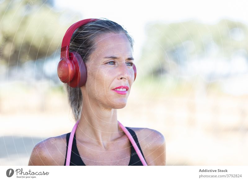 Glad sportswoman with headphones standing in park positive listen fitness device content active workout music slim vitality athlete activity training sporty