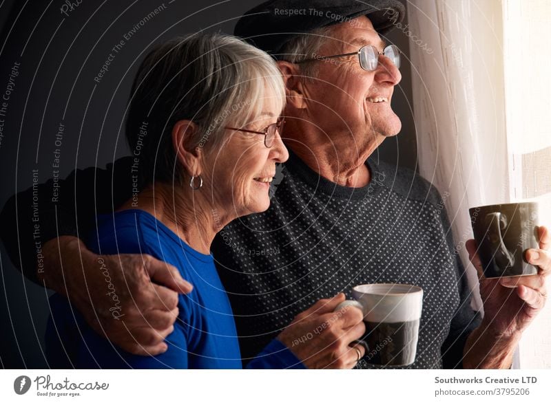 Loving Senior Couple Relaxing And Chatting Standing By Window At Home Together With Hot Drink couple senior seniors retired at home relaxing standing window