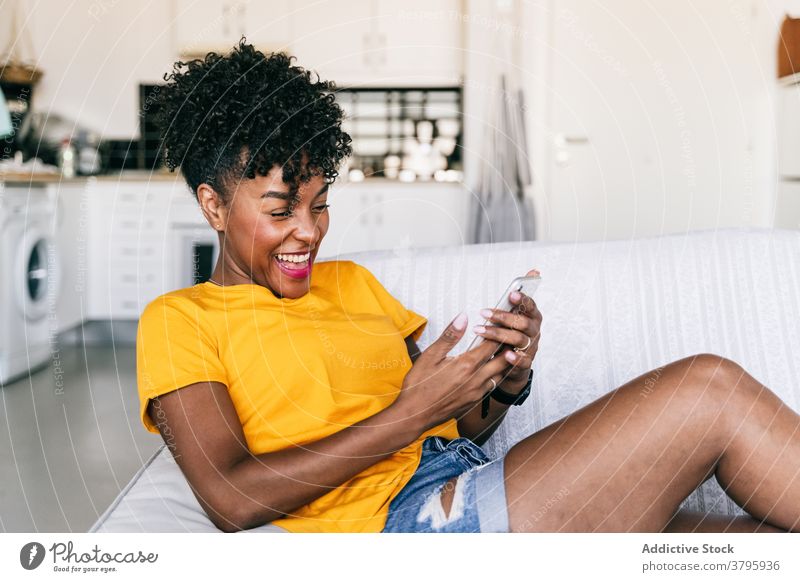 Laughing black woman using smartphone at home laugh having fun excited browsing mobile cheerful millennial young adolescent female modern device gadget