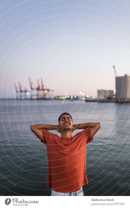 Cheerful ethnic tourist with hands behind head near ocean hand behind head smile enjoy vacation seascape sky summer twilight man traveler happy sunset water