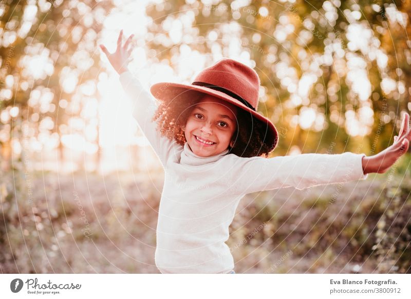 portrait of cute afro kid girl wearing a hat at sunset during golden hour, autumn season, beautiful trees background nature outdoors brown leaves