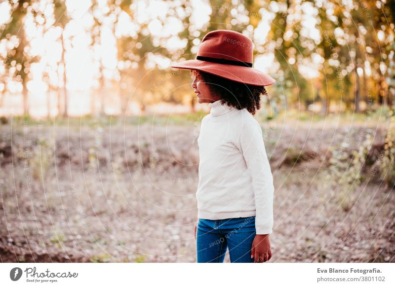 portrait of cute afro kid girl wearing a hat at sunset during golden hour, autumn season, beautiful trees background nature outdoors brown leaves