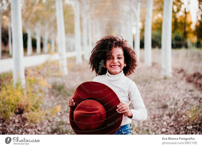 portrait of cute afro kid girl holding a hat at sunset during golden hour, autumn season, beautiful trees background nature outdoors brown leaves