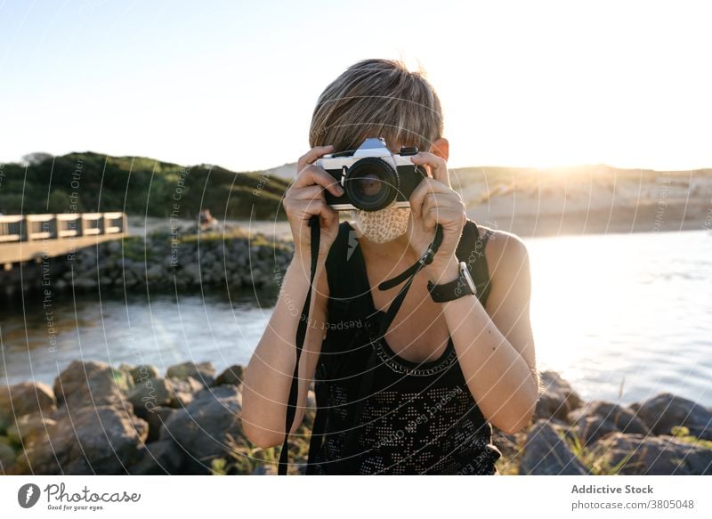 Focused young woman with photo camera relaxing on rocky seashore at sunset coast tourist peaceful admire wanderlust trip holiday vacation female short hair