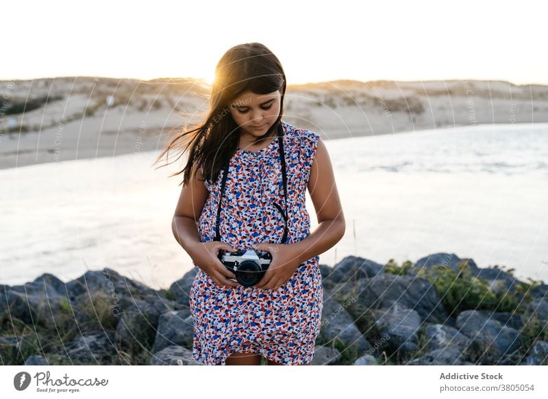 Anonymous girl shooting sea on photo camera during summer vacation woman take photo tourist sunset style holiday shore rocky travel female young dress trendy