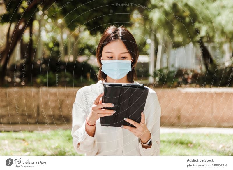 Young ethnic woman working remotely on tablet in park attentive new normal coronavirus job busy gadget using browsing female young asian formal mask covid 19