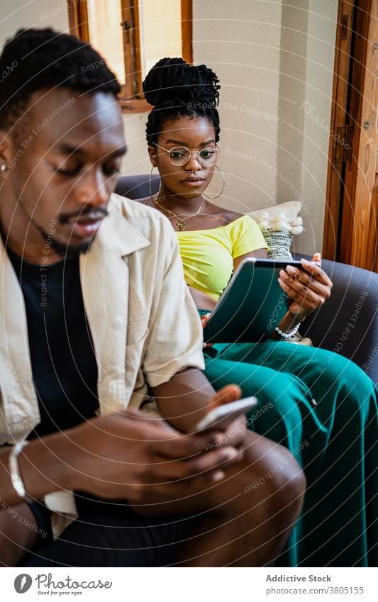 Young ethnic couple using gadgets while relaxing on couch at home sofa together relationship tablet smartphone addict connection browsing online internet