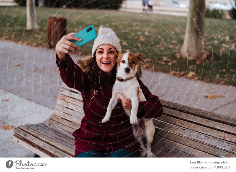 woman sitting on a bench, using mobile phone in a park with her adorable jack russell dog. Lifestyle outdoors autumn brick wall casual clothing caucasian