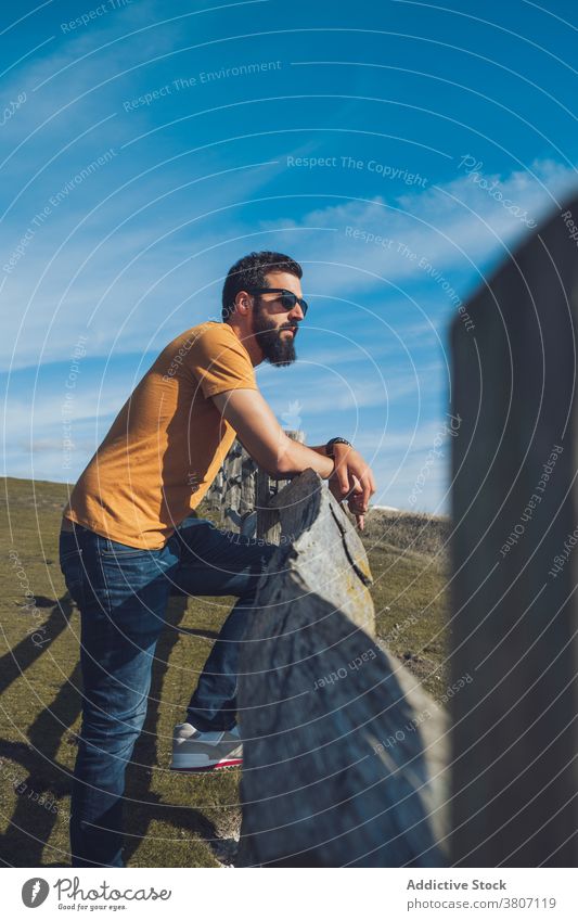 Confident young male traveler resting on grassy hill man relax admire nature confident fence wanderlust calm serene lean on ethnic casual outfit sunglasses