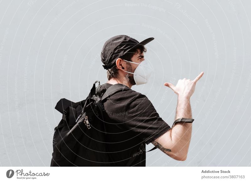 Anonymous man in mask showing shaka gesture on street backpack covid 19 self isolation respiratory wall call demonstrate rucksack trendy style apparel