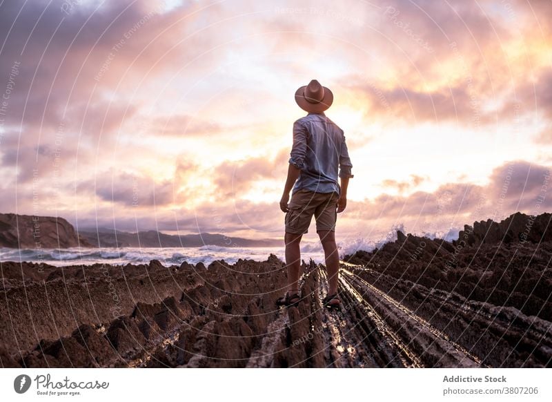 Unrecognizable tourist in straw hat near ocean at sunset traveler freedom walk shore sundown sky cloudy rocky man sandy coast atmosphere magic dramatic shiny