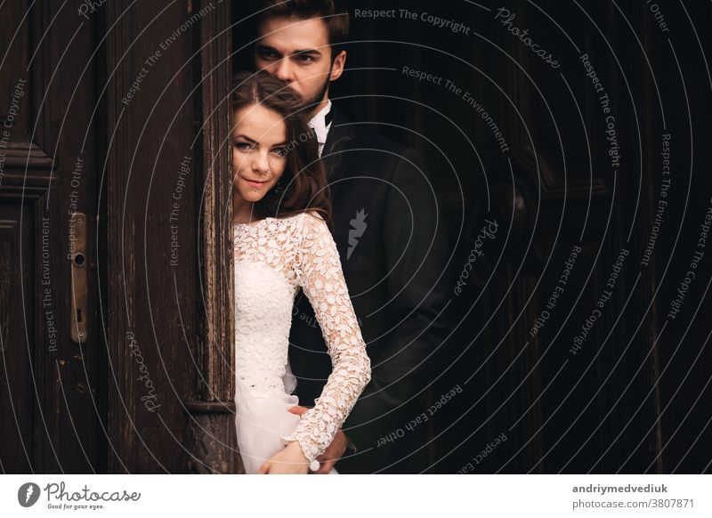 Beautiful newlyweds hugging near the ancient door. Wedding portrait of a stylish groom and a young bride near old house in in a European town embrace beautiful