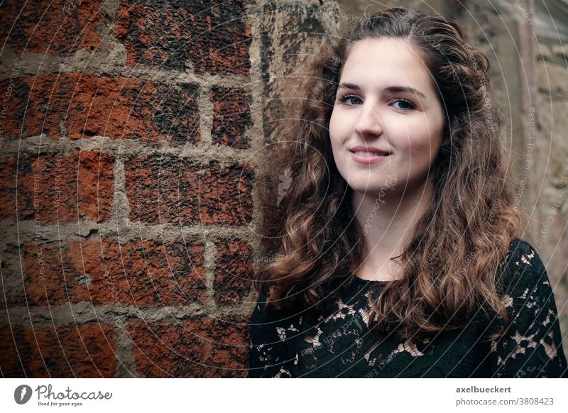 happy smiling girl leaning against old brick wall woman outdoor real authentic people looking young lady looking at camera portrait curly brunette hair