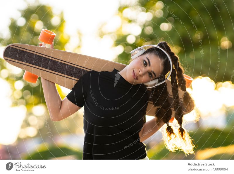 Cheerful ethnic lady with longboard enjoying music in headphones in park woman listen skate park happy active cheerful trendy tree training modern female young