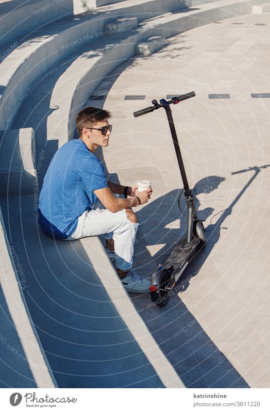 Young Caucasian man sitting outdoor near electric scooter and drinking coffee to-go cup to go Student close up Guy Male Ecology City blue grey Urban Modern