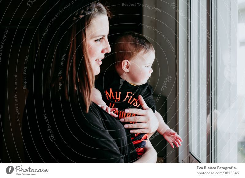 young mother and baby girl at home looking by the window. Baby girl wearing Halloween clothes. Family lifestyle indoors love together family childhood