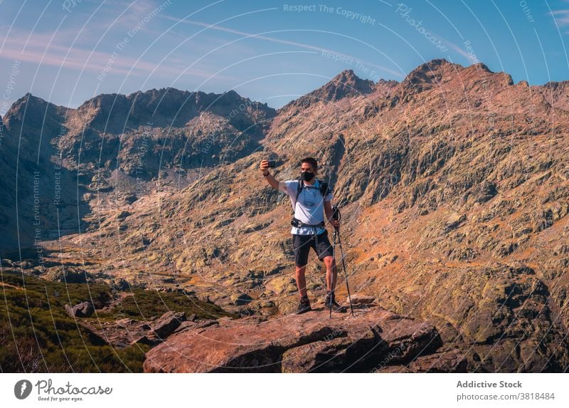 Anonymous tourist taking selfie on smartphone in mountains highland nature vacation mask covid19 using gadget cellphone trekking pole blue sky cloudy traveler