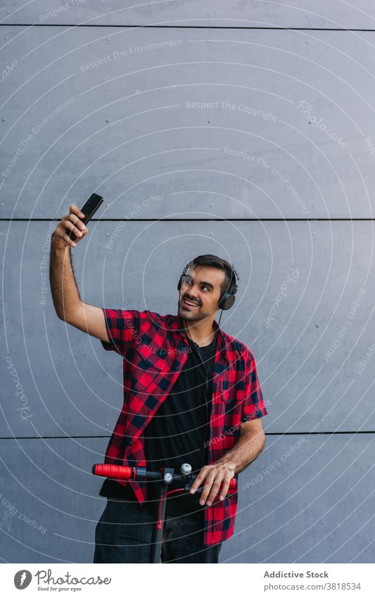 Cheerful man on electric scooter taking selfie in city hipster style checkered shirt headphones entertain male smartphone self portrait modern urban music