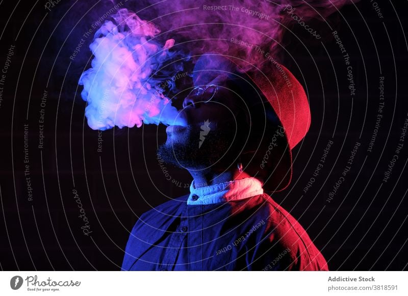 Ethnic man smoking in studio with neon lights smoke vape steam style illuminate dark male ethnic black african american sunglasses hat trendy hipster cool