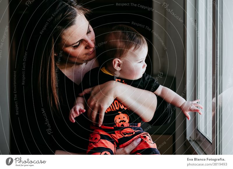 young mother and baby girl at home looking by the window. Baby girl wearing Halloween clothes. Family lifestyle indoors love together family childhood