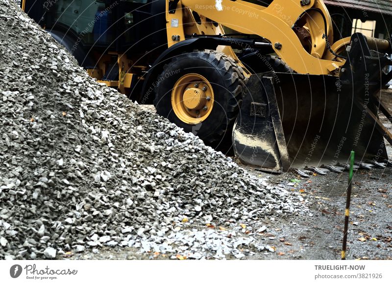 Road construction and landscaping with a heavy yellow wheel loader, thick black tyres and loading shovel next to a grey heap of coarse-grained sand or gravel and a thin iron bar stuck crooked in the ground