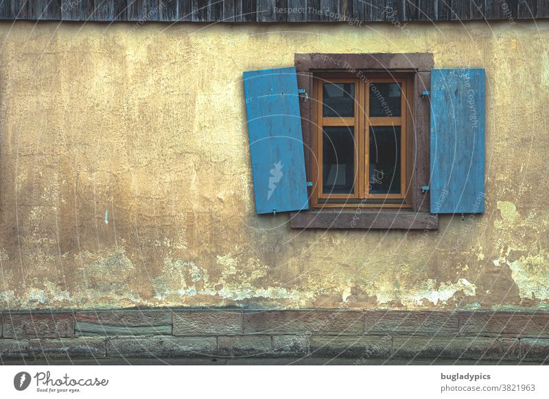 Yellow/ terracotta/ beige façade with a brown sash bar window and blue/ turquoise shutters. One of the shutters hangs crooked. Facade Shutter Lattice window