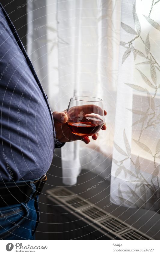 Overweight man with a glass of cognac standing at a window Man Cognac drinking glass Hand stop Window Observe Loneliness at home stay at home Fat coronavirus