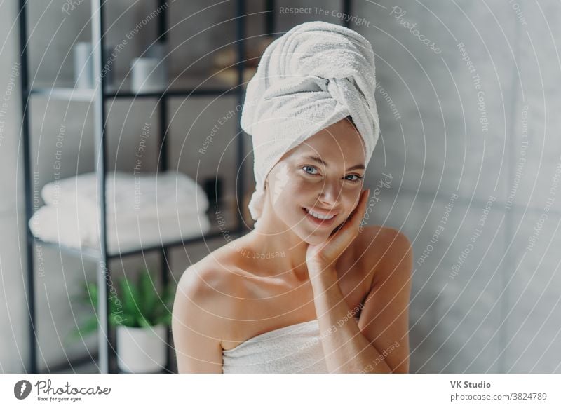 Beautiful European woman with makeup touches skin, has minimal makeup, has healthy glowing skin, wrapped in bath towel, enjoys rest at home. Spa woman poses in bathroom. Beauty, wellness concept