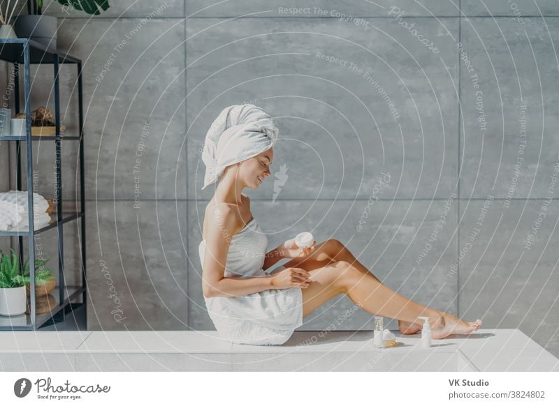 Horizontal shot of relaxed young European woman applies moisturizing body cream on legs, wrapped in bath towel, has tender smile, healthy refreshed skin after taking bath, poses in cozy bathroom.