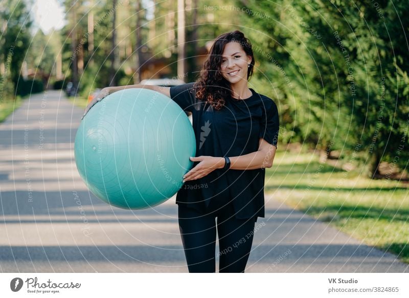 Outdoor shot of brunette woman poses with big fitball, dressed in sportclothes, has training in open air, walks on road, poses near forest. Fitness trainer going to show masterclass for pregnant women