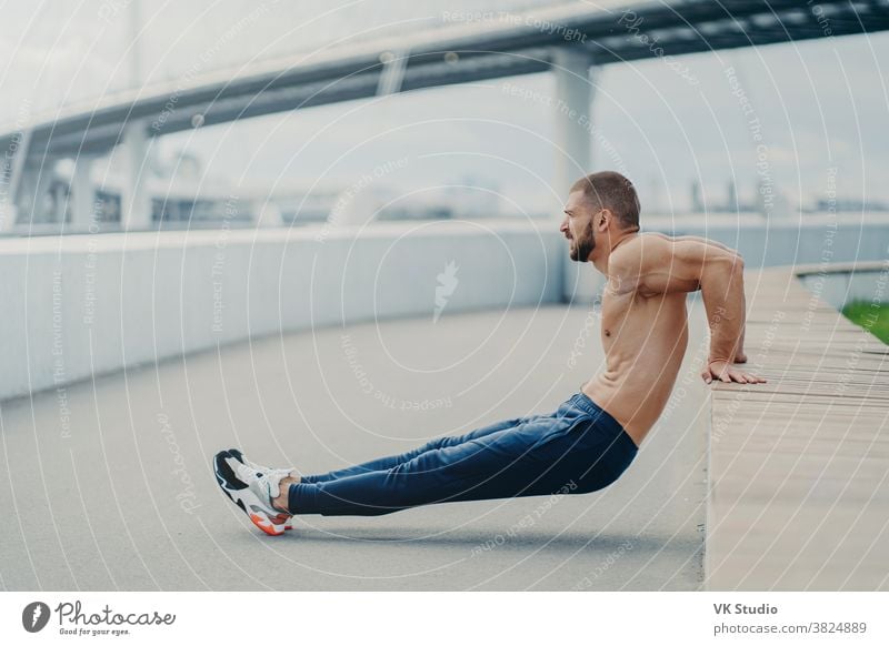 Sideways shot of muscular man does reverse push up exercise, trains arms, keeps perfect body and good physical shape, has active workout outdoor, wears sport trousers and sneakers, poses on bridge