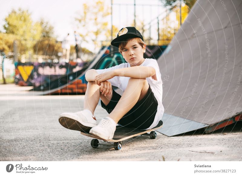 A beautiful teenager is sitting on a skateboard in a special area of the Park. A boy is resting after riding in a skatepark. Active rest in the fresh air kids