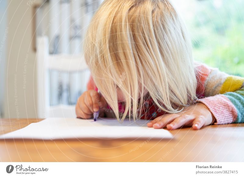 4 year old blonde girl with a brightly colored striped sweater sits at a wooden table and draws with a purple pencil on a piece of white paper child's hand