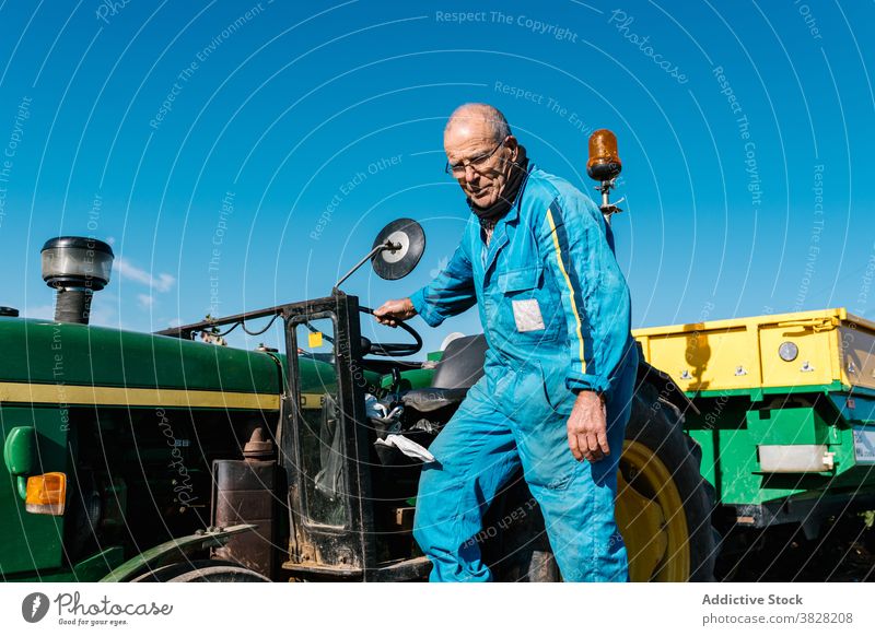 Elderly male worker near garden tractor under blue sky workman uniform style old vehicle farm bright eyeglasses countryside transport summer nature eyewear