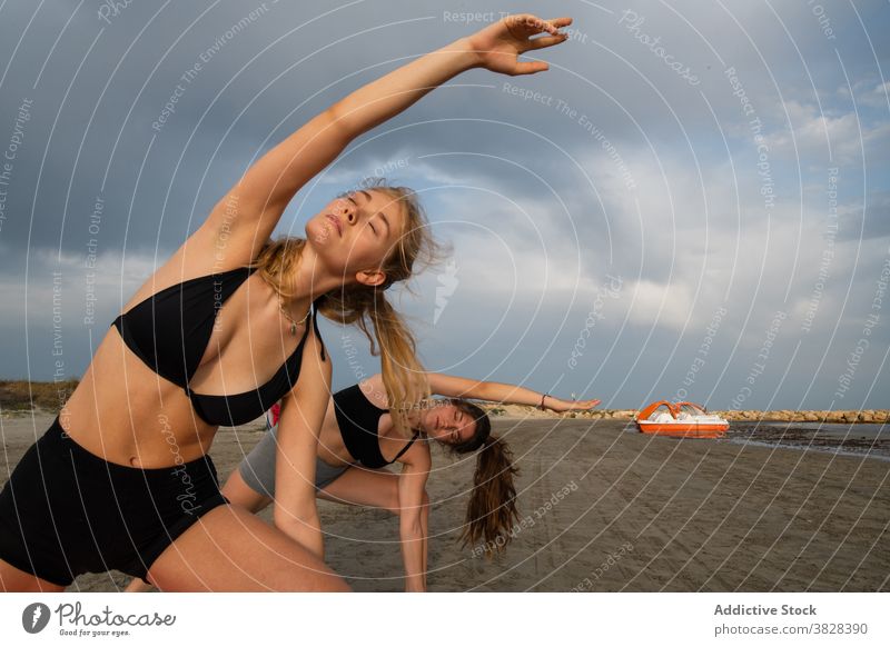 Young friends doing yoga on sunny beach serious flexible practice sandy coast rippling river gloomy day cloudy healthy fit slim concentrate balance wellness