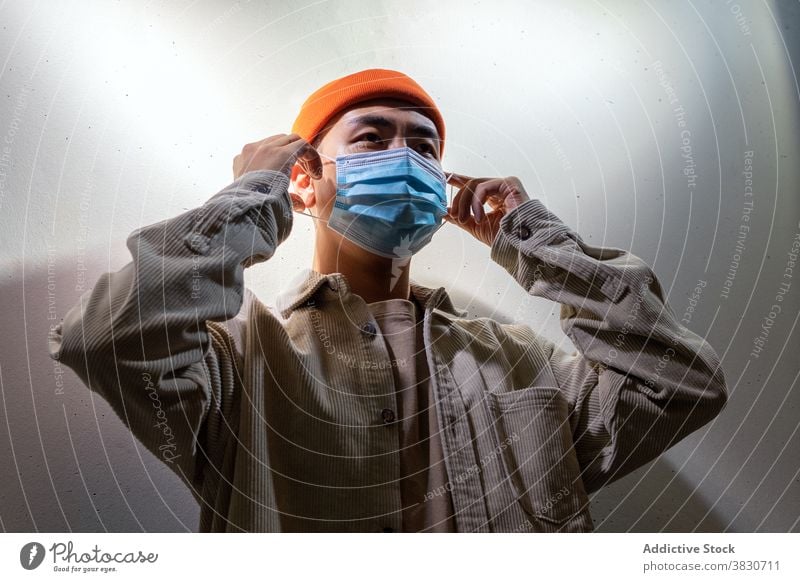 Serious Asian man in face mask standing in gray corridor respirator concentrate thoughtful pensive calm cover mouth coronavirus covid 19 ponder focus room