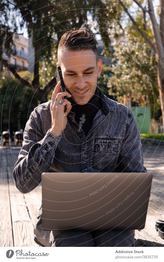 Cheerful young man working on laptop and talking on phone using typing cheerful internet freelance browsing occupation male remote device modern surfing