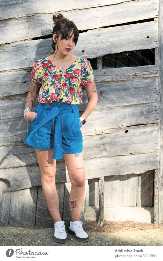 Thoughtful woman next to old wooden construction thoughtful wall style fashion dreamy exterior pensive slender female shorts hand in pocket shirt dark hair