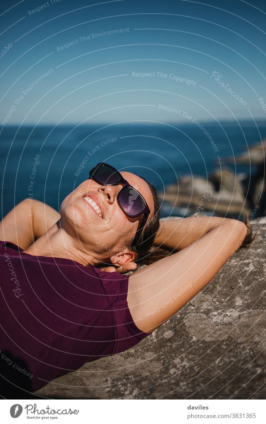 Lying woman on a rock, at the coast line with sunglasses and purple t shirt, and the sea in the background. smile waves water lying down arms ocean spain