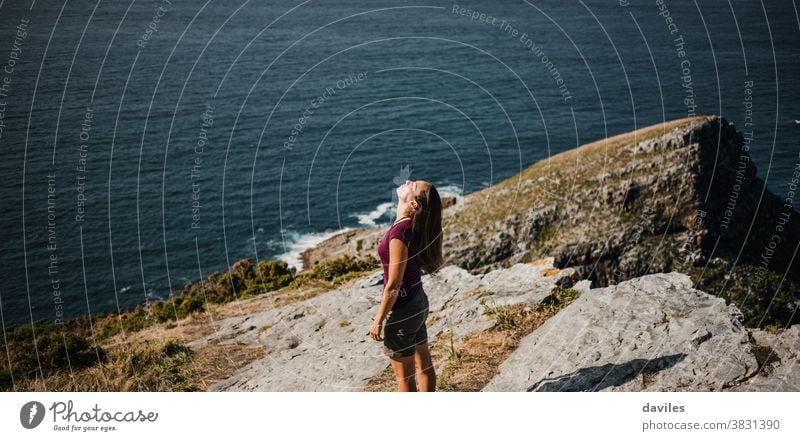 Woman standing at the coastline on the top of a cliff, with sea in the background. nice inspirational pensive sightseeing traveler elevation rock purple t shirt