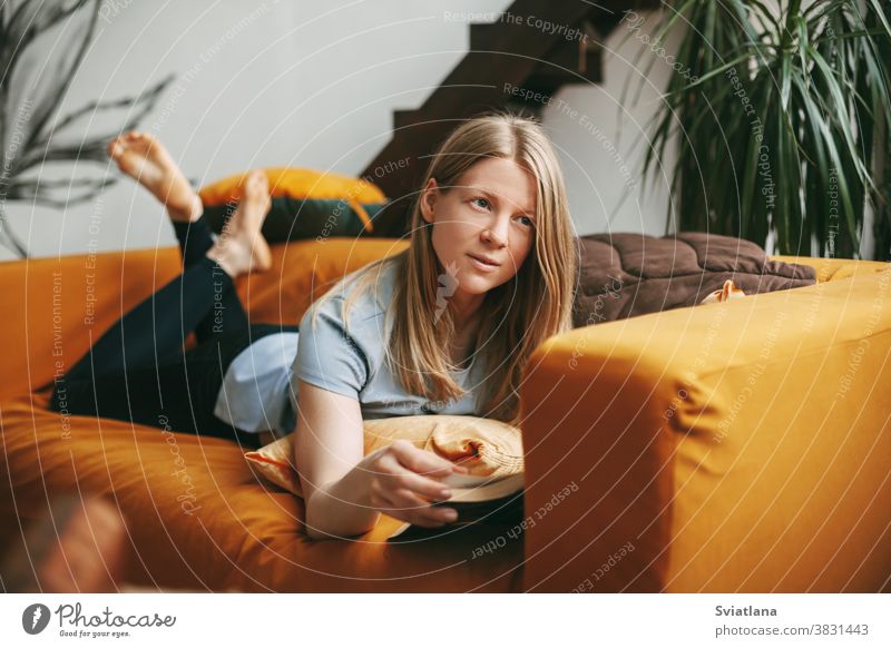 A beautiful girl is relaxing on the sofa in the living room, reading her favorite novel. Cute, thoughtful girl is lying on the couch and reading a book. happy