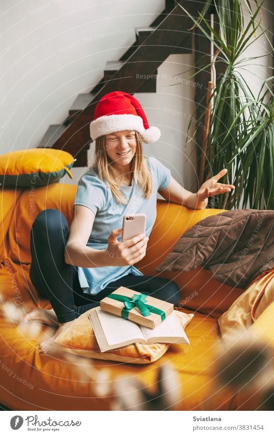 A smiling girl in a Christmas hat sits on the sofa and communicates via video link with friends. Girl congratulates friends and family on holidays christmas